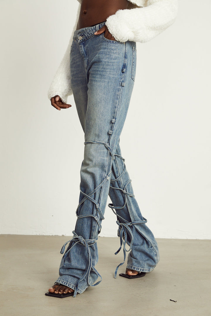 Light wash blue jeans with layered strap details and tie-up cuffs on the lower leg