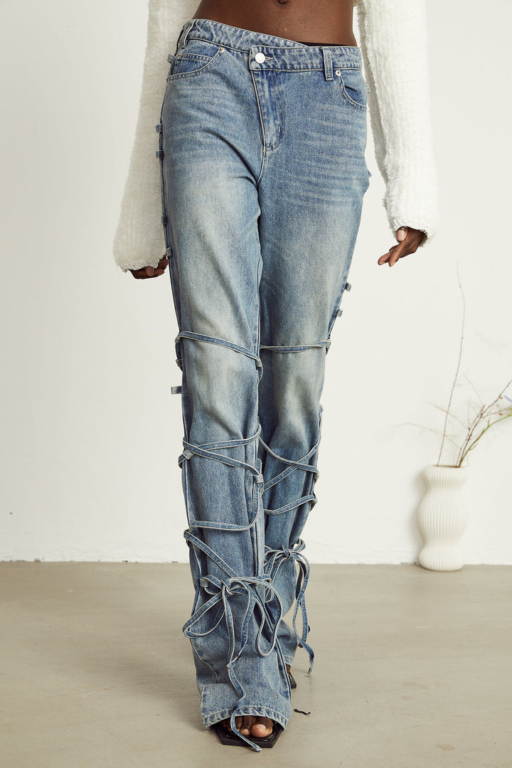 Light wash blue jeans with layered strap details and tie-up cuffs on the lower leg
