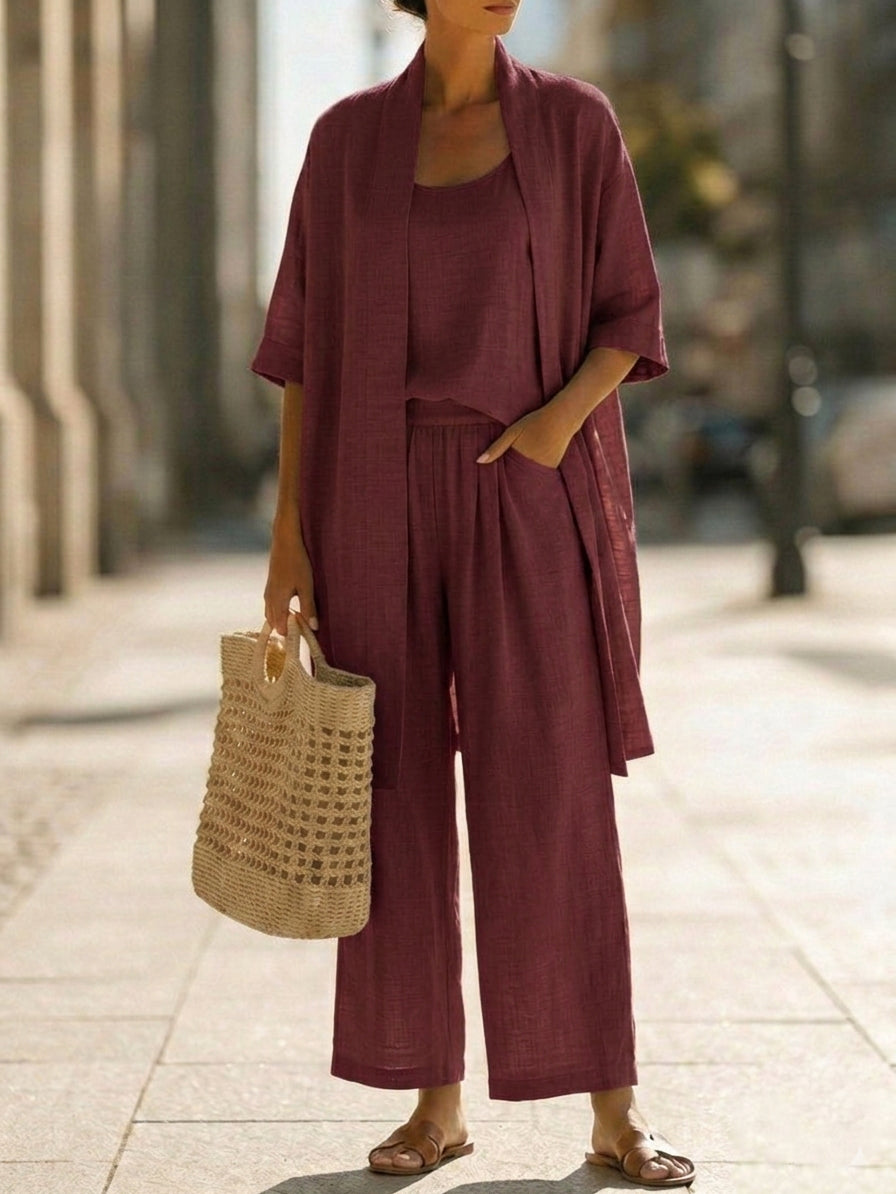 Woman wearing white three-piece set with open cardigan, scoop-neck top and wide-leg trousers, holding woven tote bag
