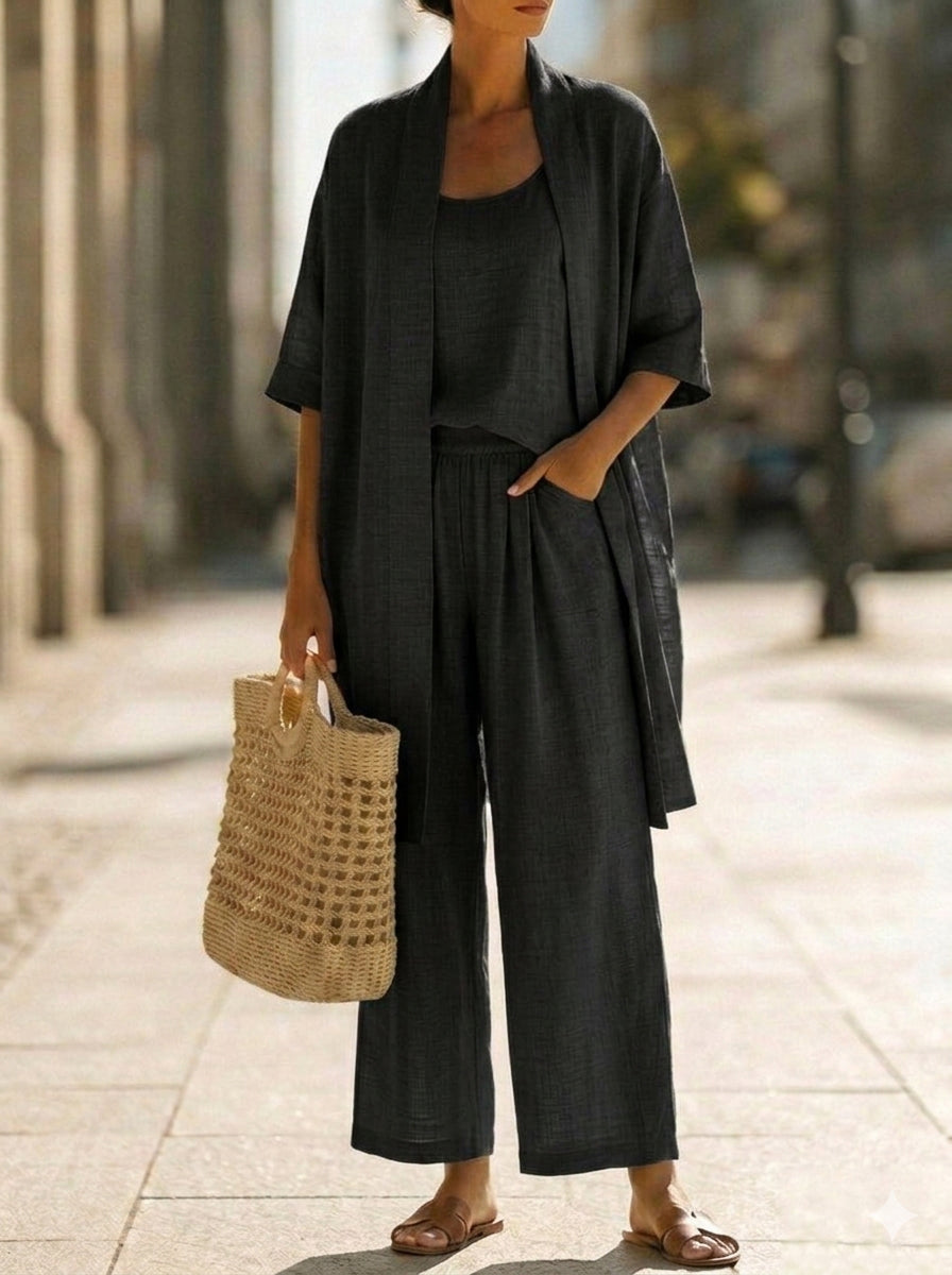 Woman wearing white three-piece set with open cardigan, scoop-neck top and wide-leg trousers, holding woven tote bag