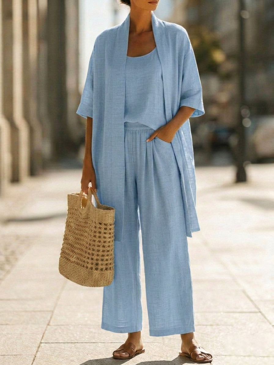 Woman wearing white three-piece set with open cardigan, scoop-neck top and wide-leg trousers, holding woven tote bag