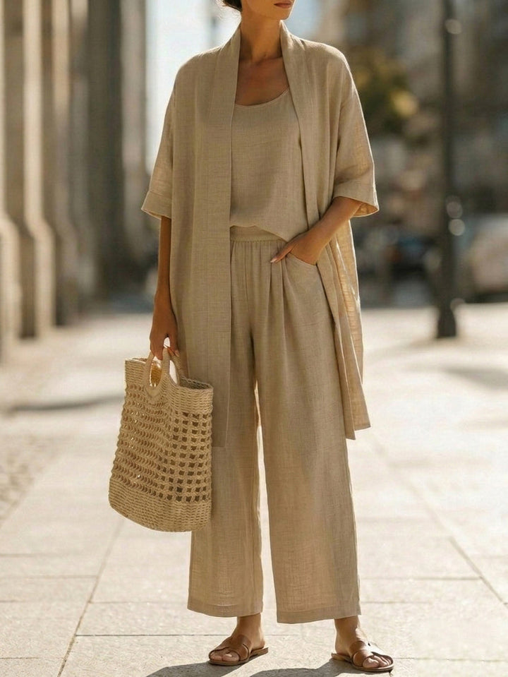 Woman wearing white three-piece set with open cardigan, scoop-neck top and wide-leg trousers, holding woven tote bag