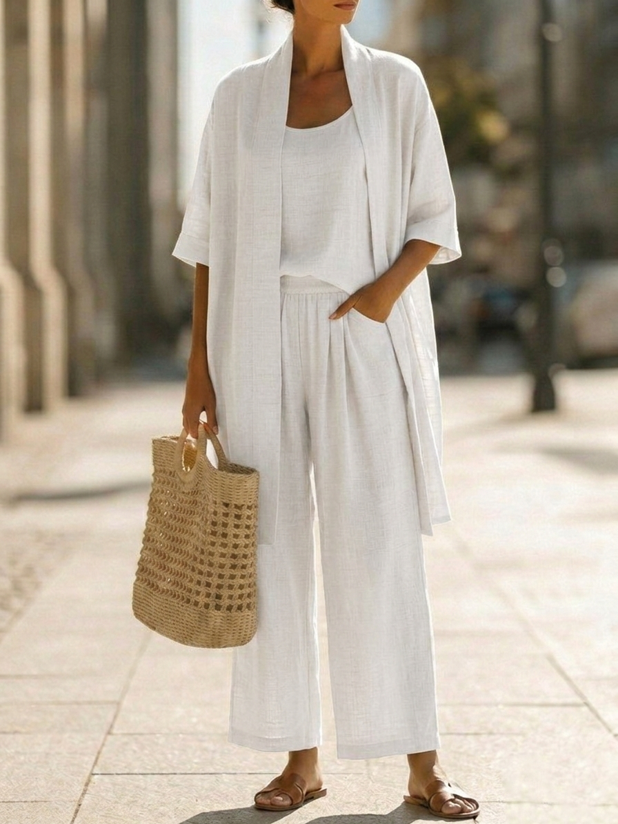 Woman wearing white three-piece set with open cardigan, scoop-neck top and wide-leg trousers, holding woven tote bag