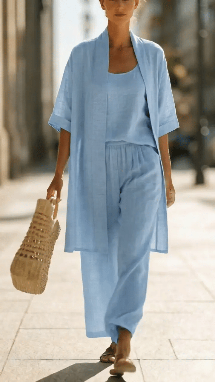 Woman wearing white three-piece set with open cardigan, scoop-neck top and wide-leg trousers, holding woven tote bag